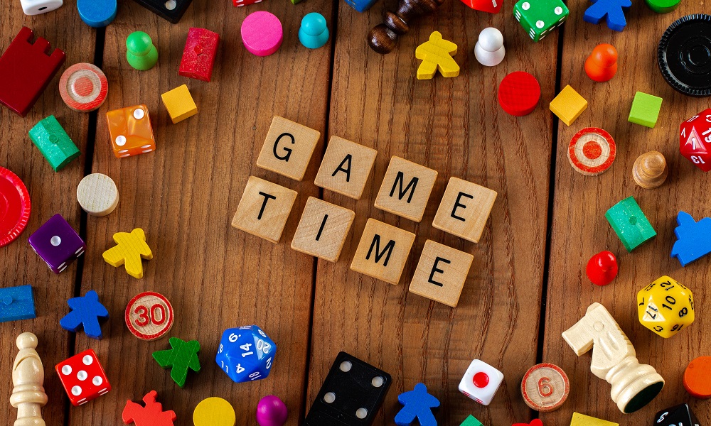 "Game Time" spelled out in wooden letter tiles. Surrounded by dice, cards, and other game pieces on a wooden background