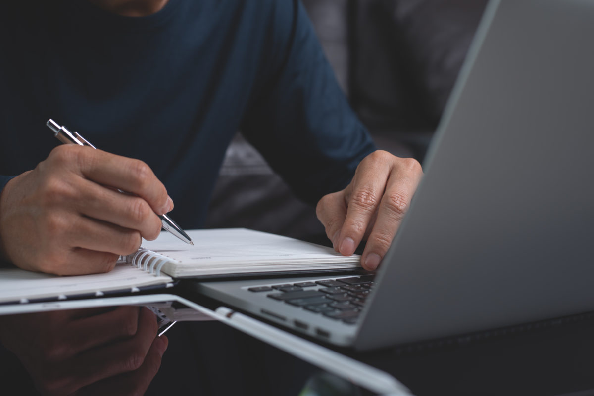 Casual business man sitting at wooden table writing note on paper notebook while working on laptop computer in office. Student attending online course and doing homework. Online education,e-learning concept